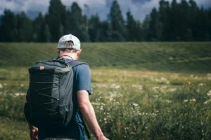 A man in a cap walking through a green field with a dark daypack on his back