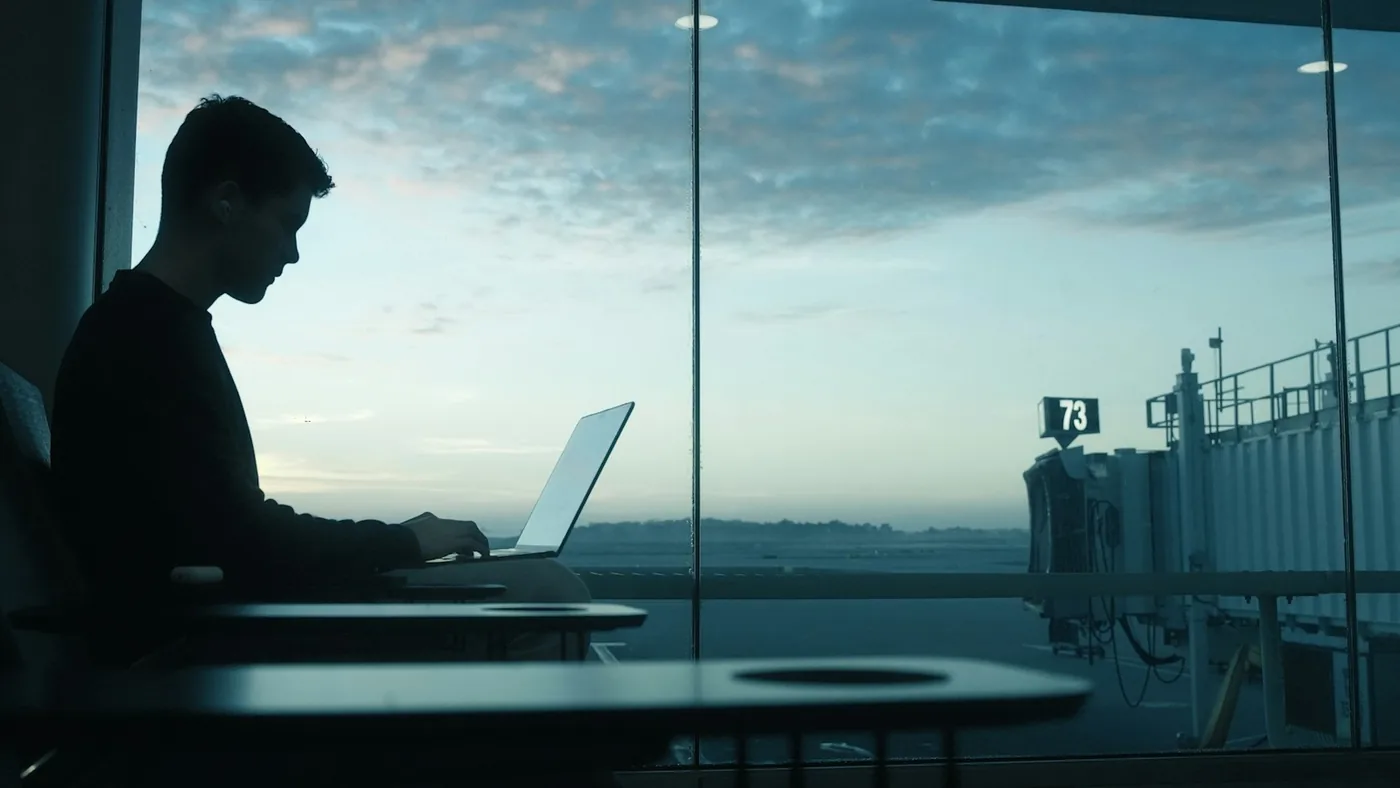 Man working on a laptop by an airport window at sunrise