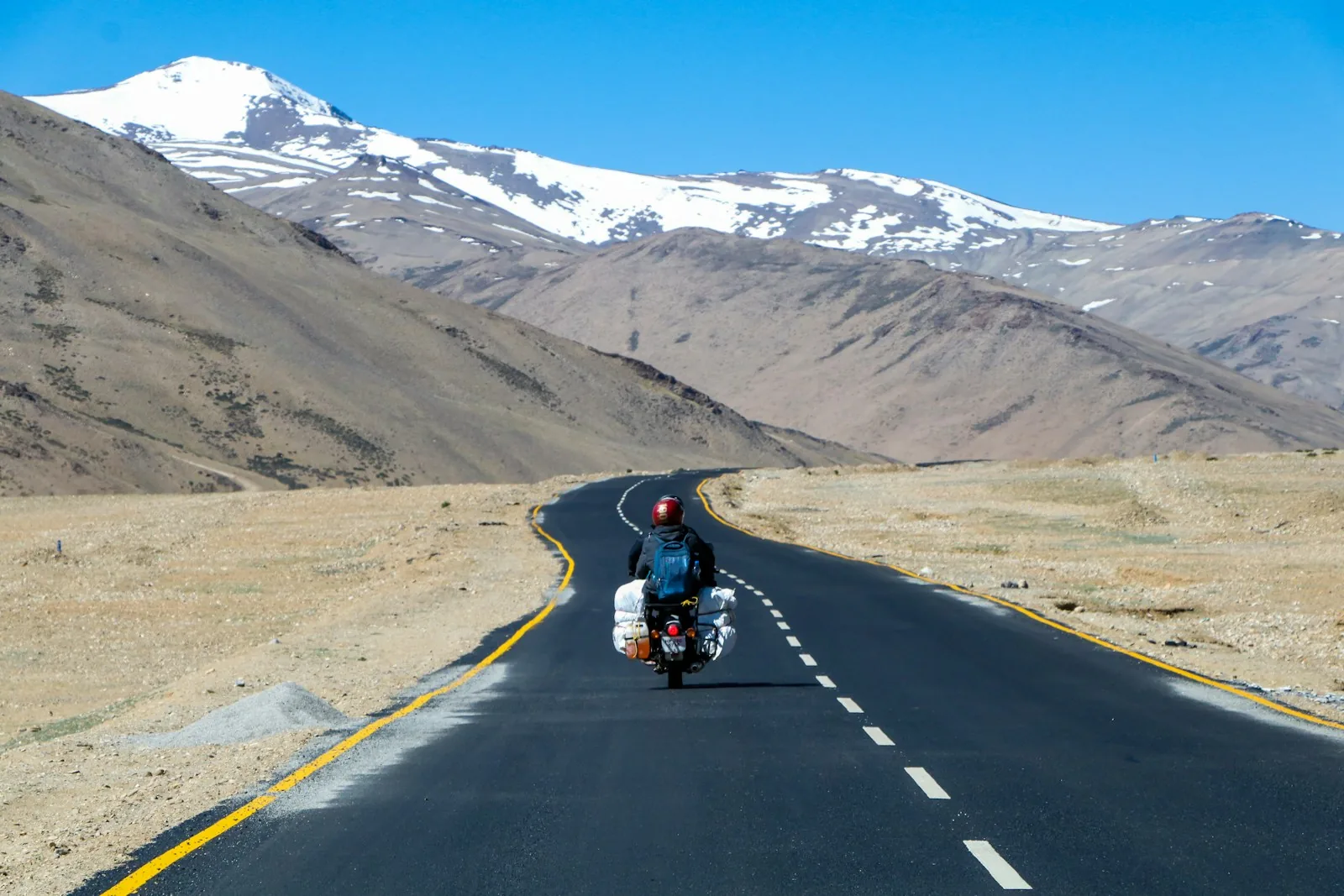 A rider on a loaded motorcycle on an empty mountain road below snowy peaks