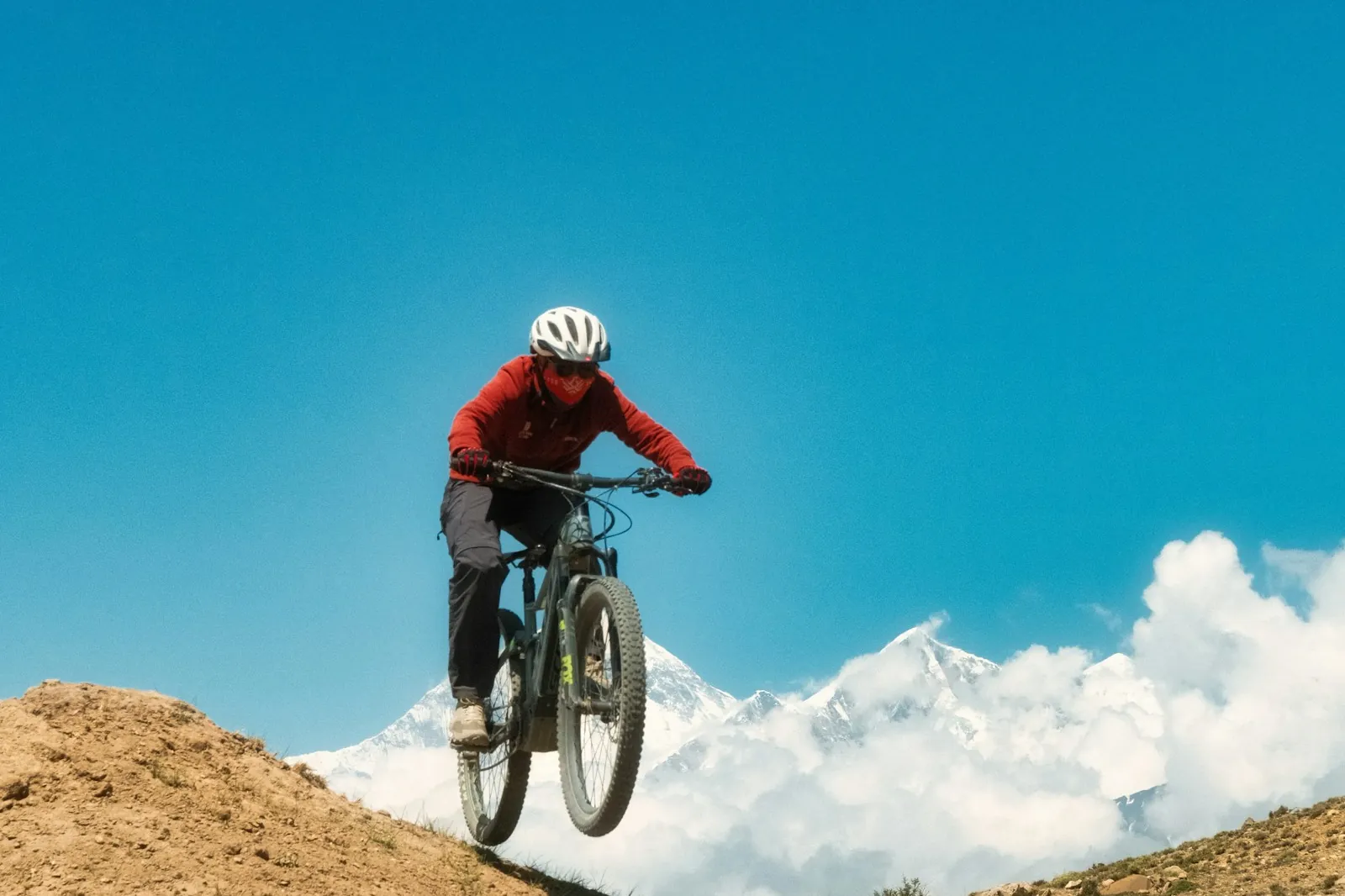 Un ciclista de montaña con chaqueta roja saltando desde una rampa de tierra, con picos nevados y nubes al fondo