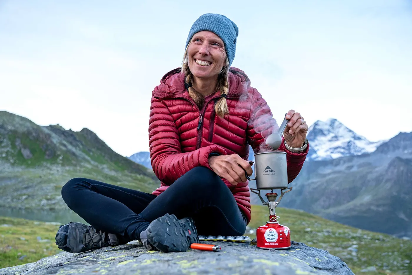 Lächelnder Wanderer in roter Jacke rührt in einem Topf auf einem MSR PocketRocket 2 auf einem Felsen, im Hintergrund schneebedeckte Gipfel