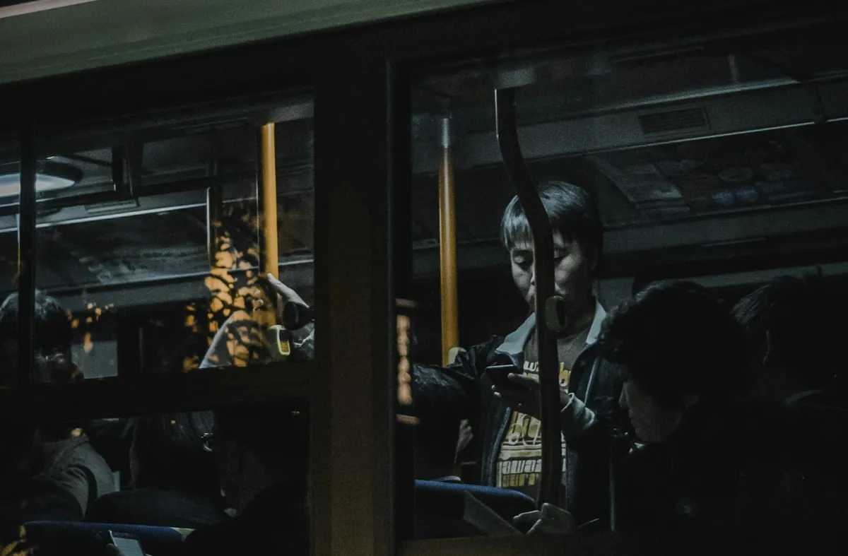 Passenger looking at a phone inside a bus at night, seen through the window