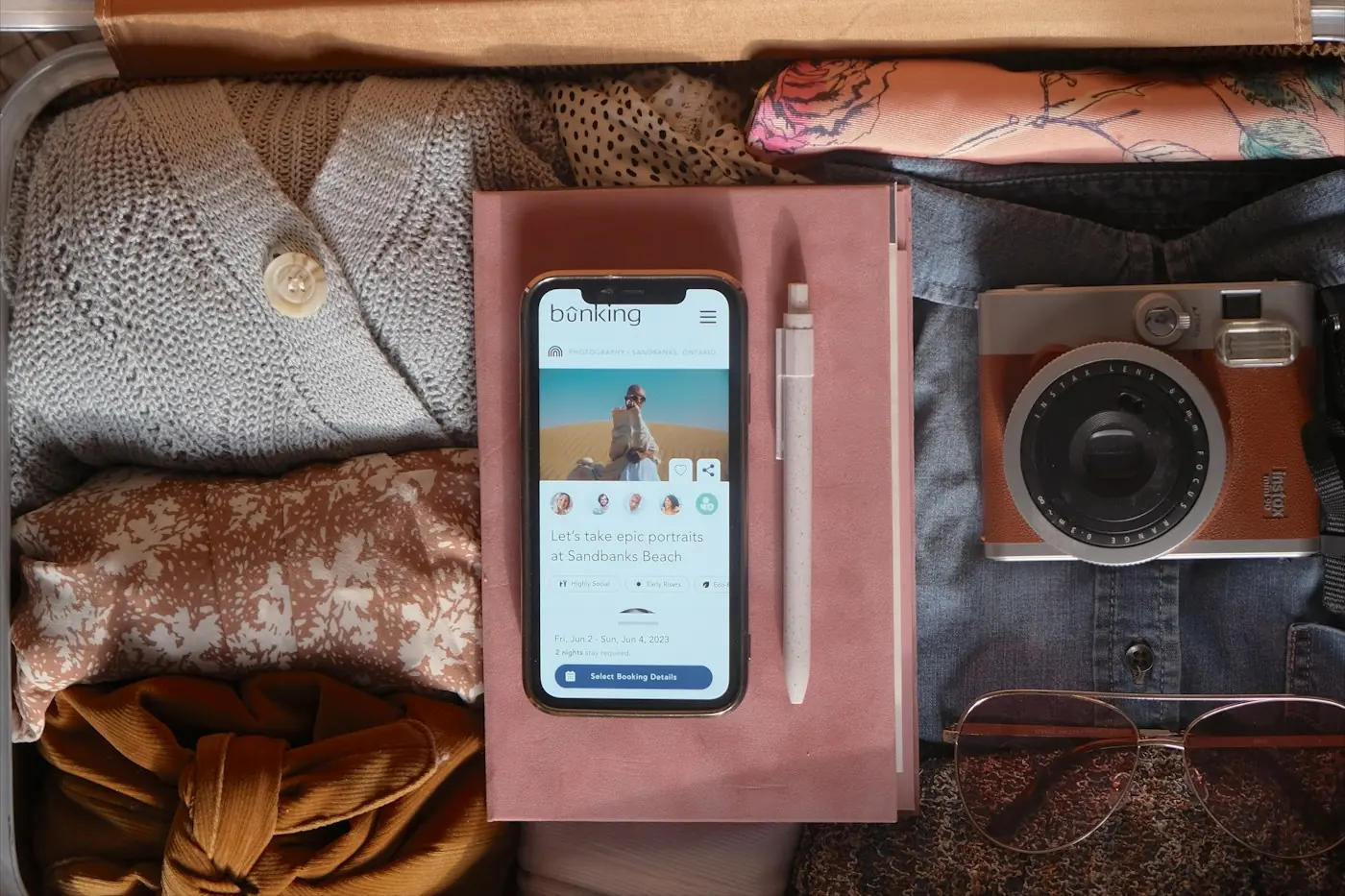 Open suitcase seen from above with folded clothes, a phone on a notebook, a camera and sunglasses