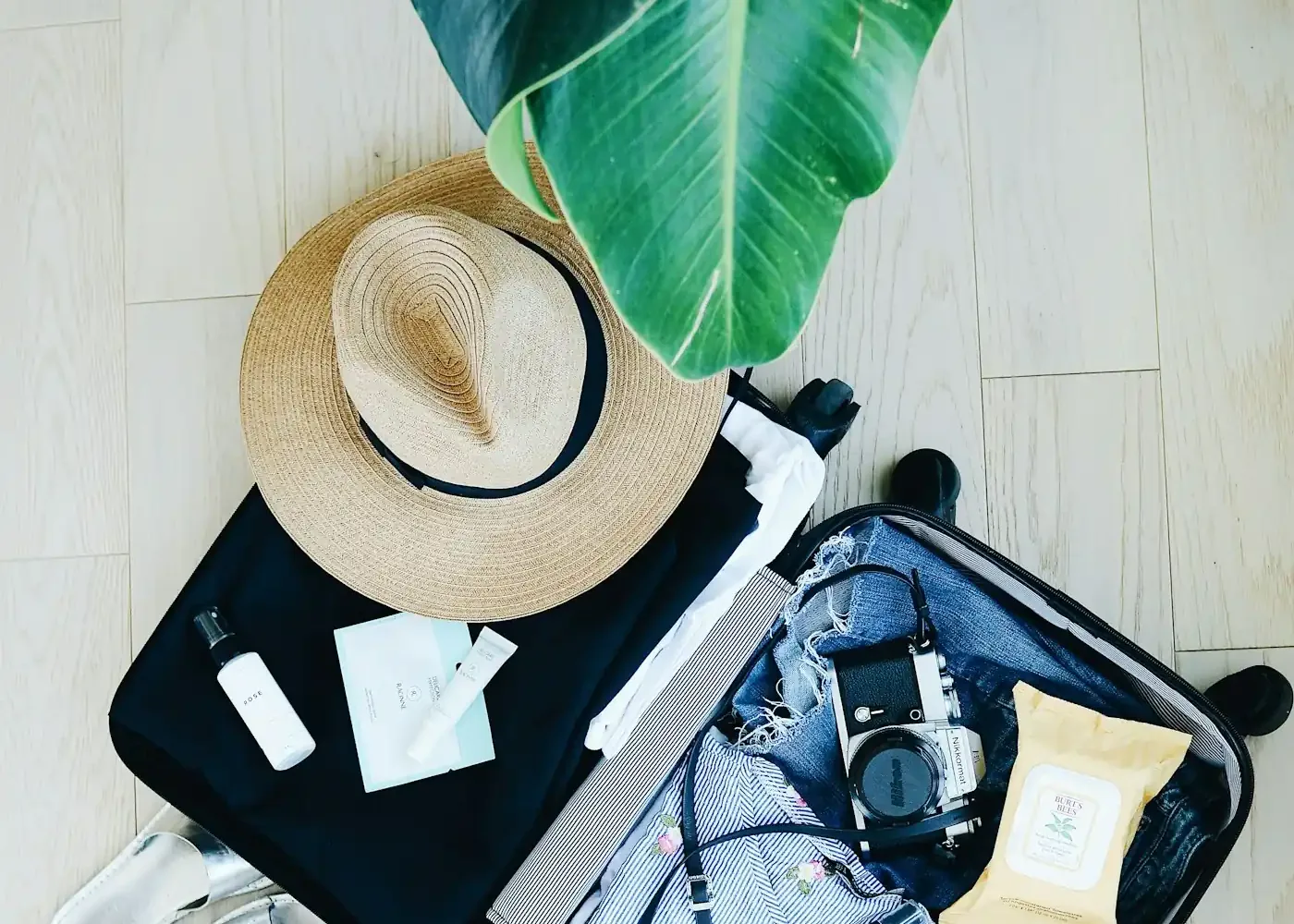 Open suitcase on a light floor with a straw hat, a camera, folded clothes and toiletries next to a banana leaf