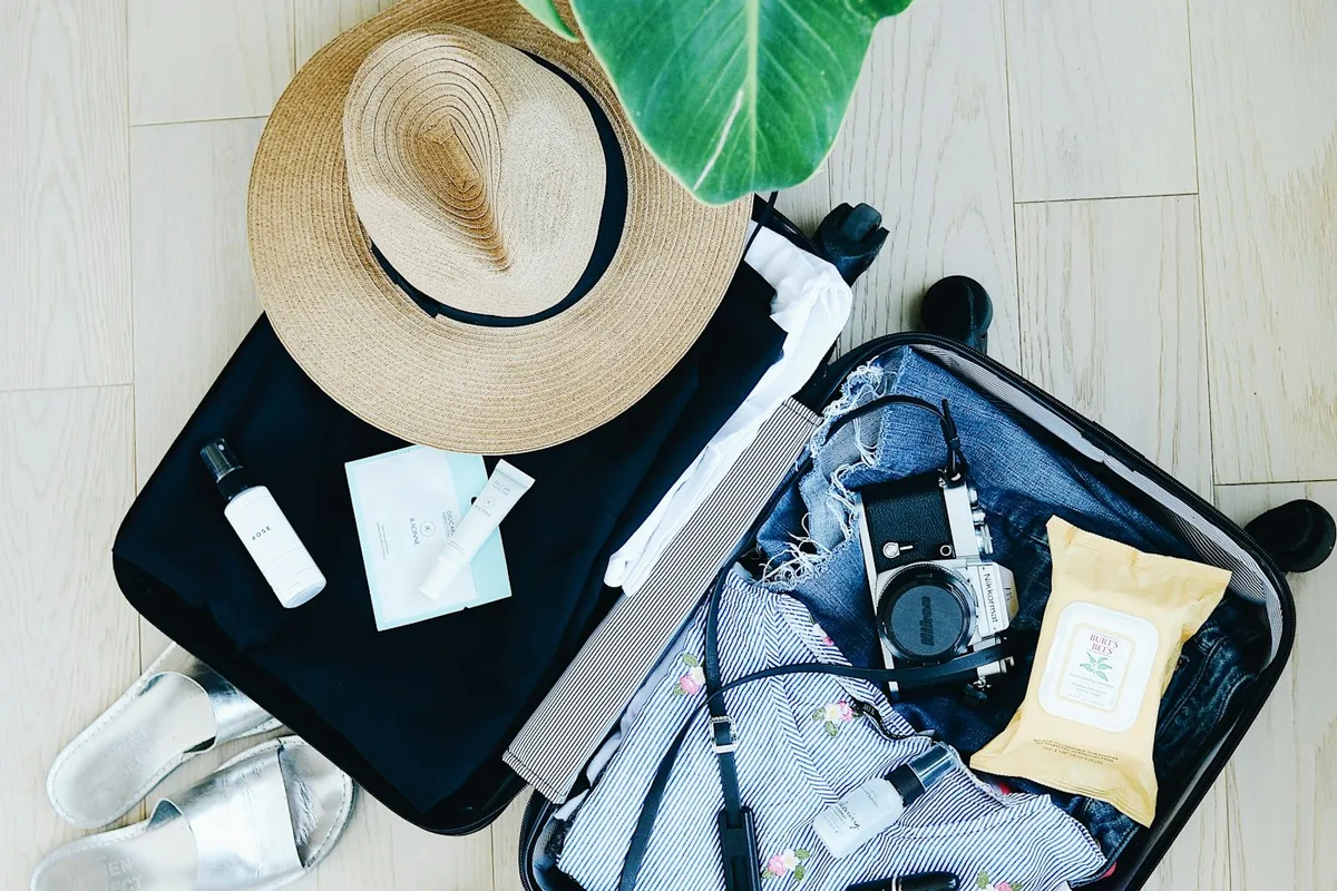 An open suitcase with folded clothes, a straw hat and travel essentials next to a plant