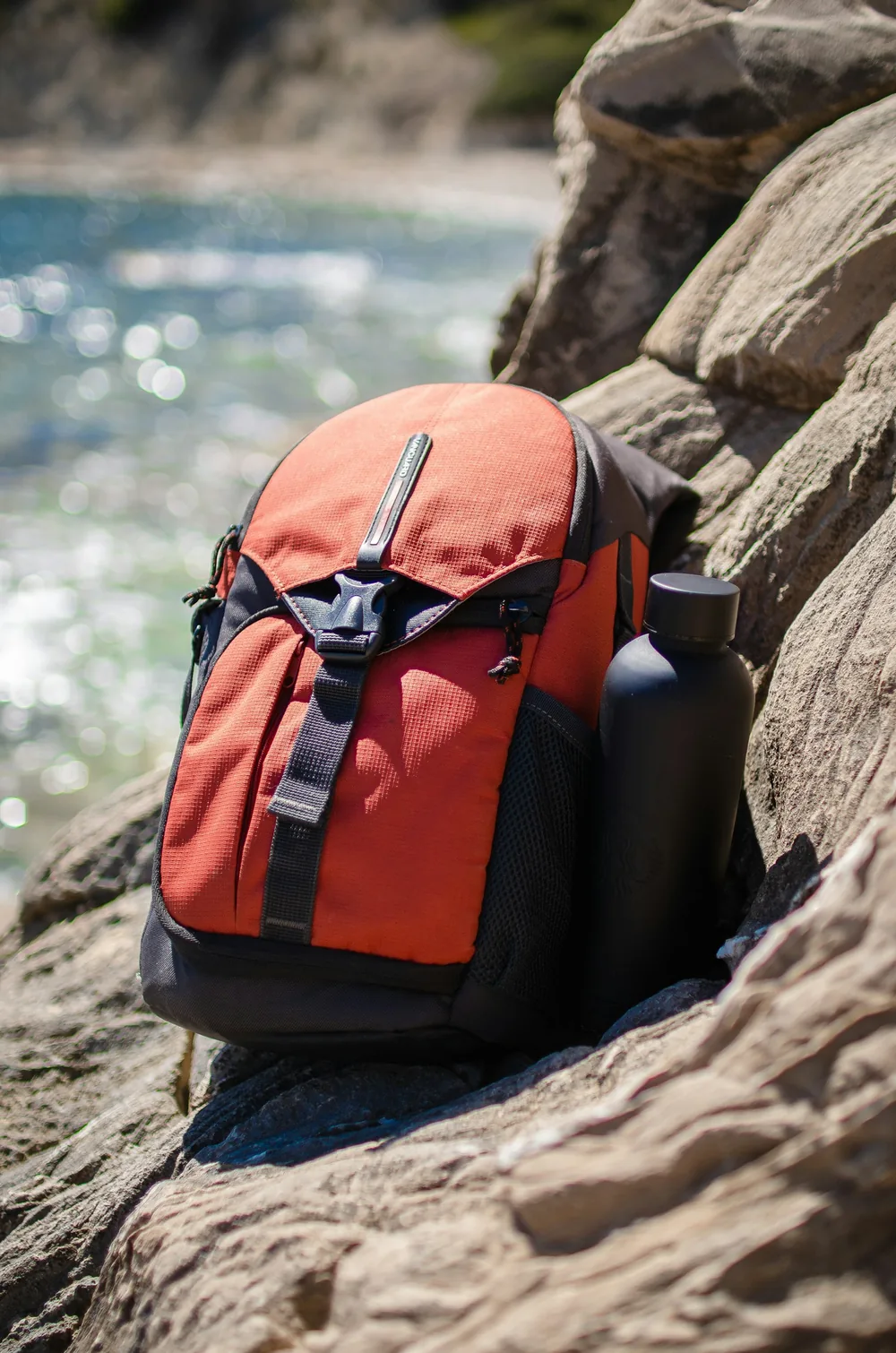 Una mochila de día naranja con un bolsillo lateral para botella, apoyada sobre rocas junto al mar