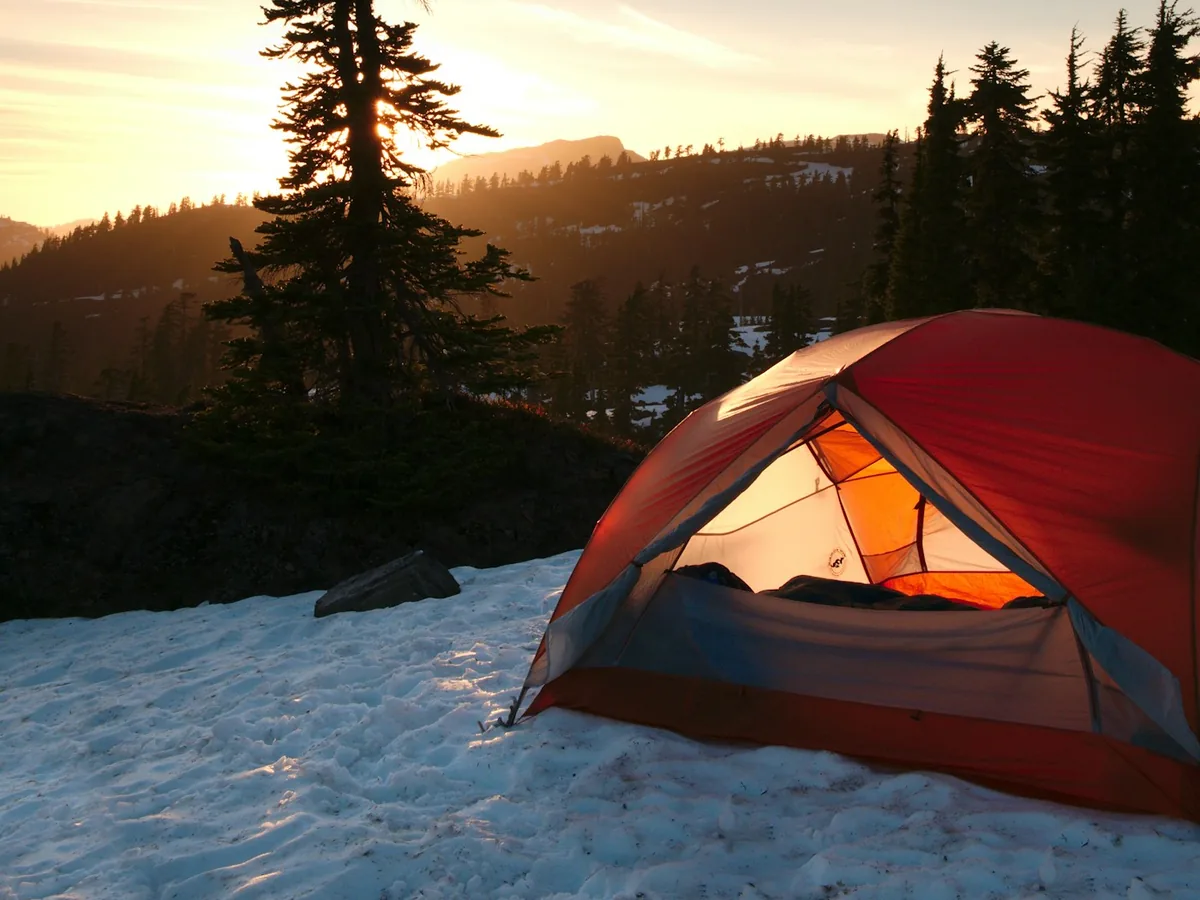 Una tienda de campaña tipo cúpula roja y naranja montada sobre la nieve al amanecer junto a un pino