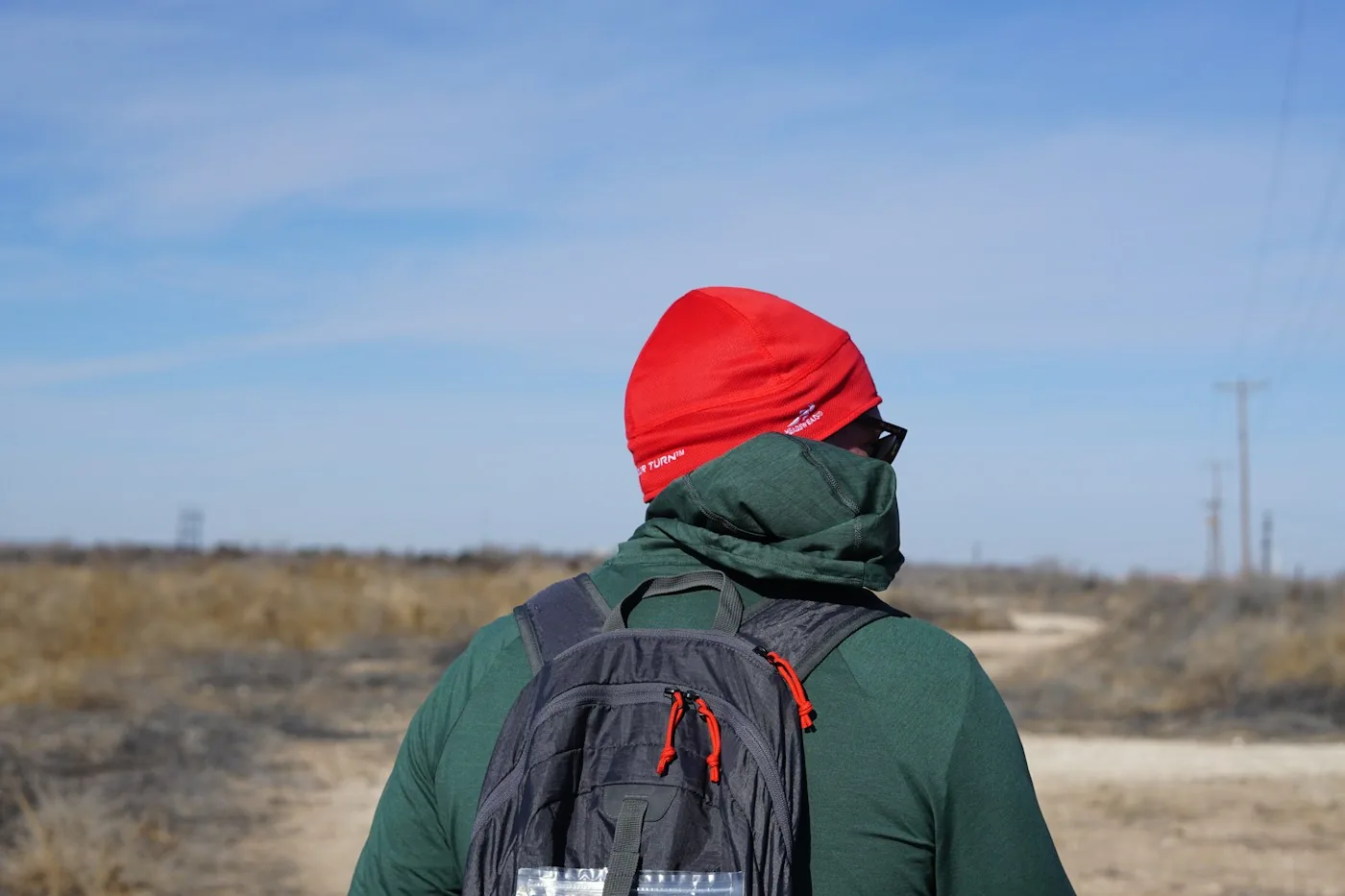 Traveler in a green jacket and red hat with a grey backpack on a dirt road