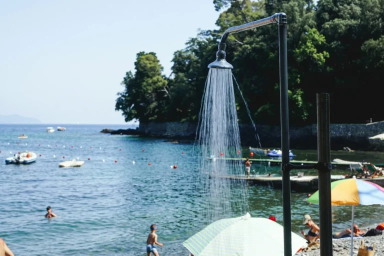 An outdoor shower spraying water on a beach with a bay and swimmers behind it