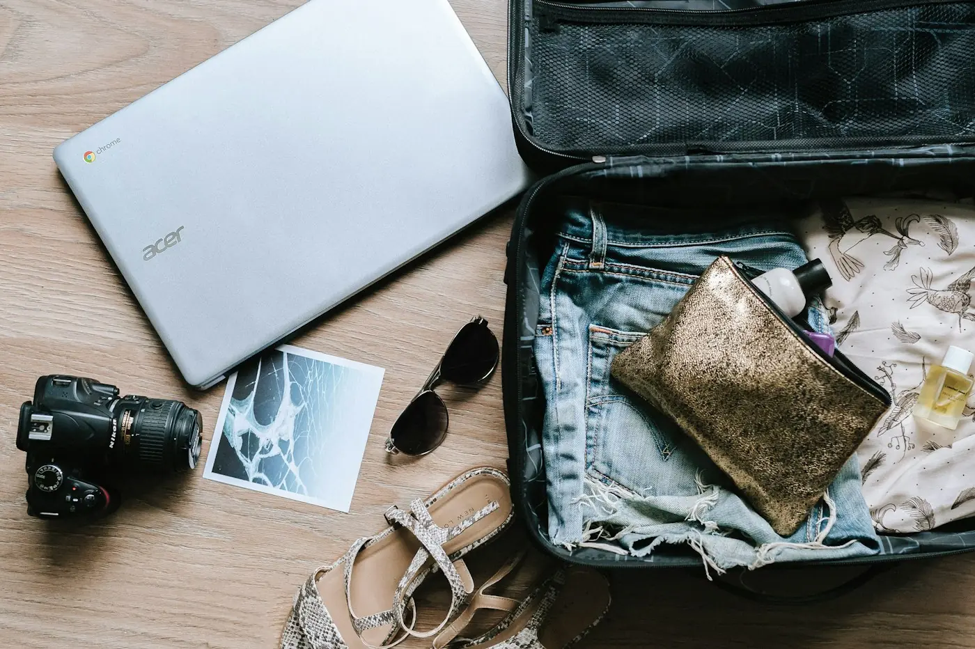 Portátil, cámara analógica, gafas de sol y una bolsa abierta y hecha sobre un suelo de madera