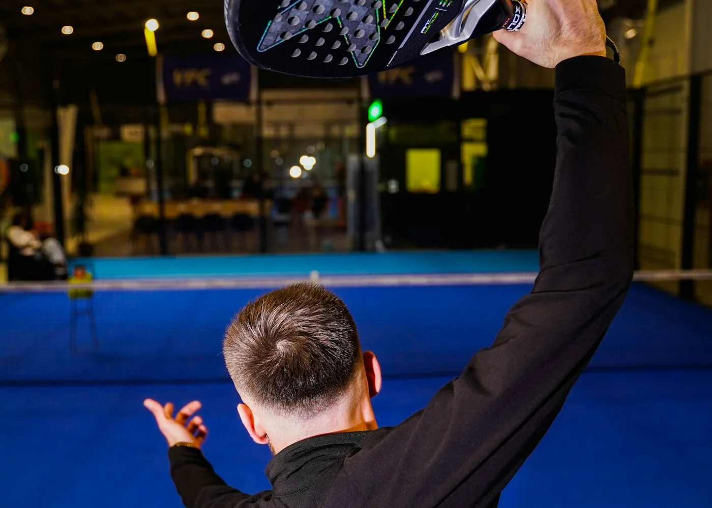 Jugador de pádel visto de espaldas con el brazo que sostiene la pala en alto en una pista azul interior