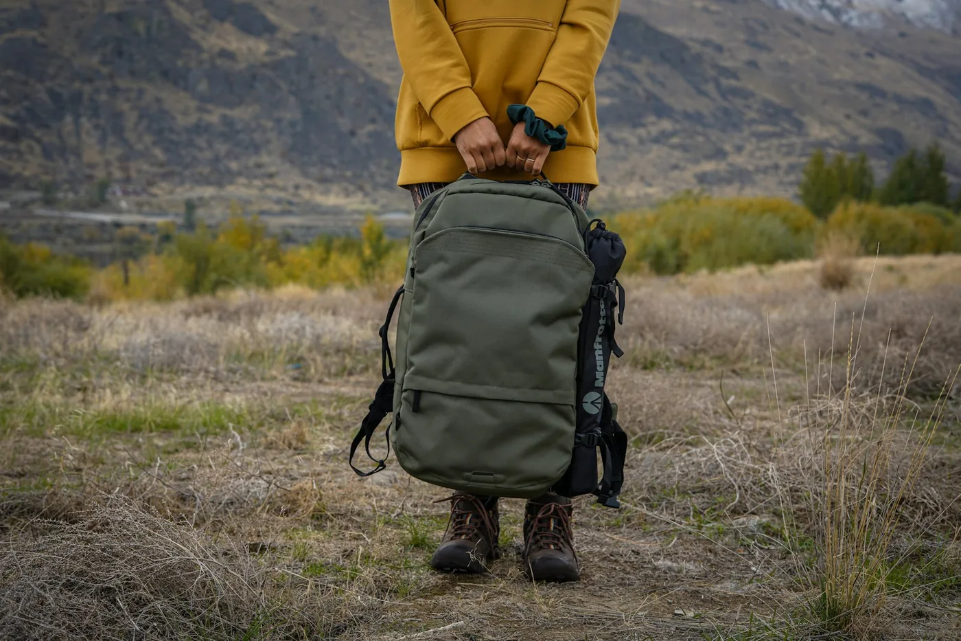Una persona sosteniendo una mochila de día verde frente a las piernas, en un campo de hierba seca con montañas al fondo