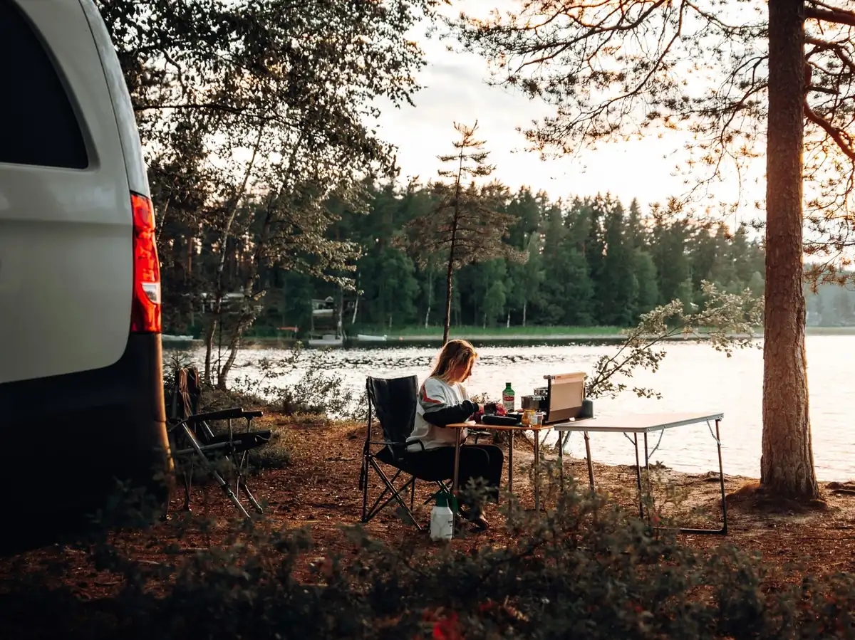 Persona sentada en una pequeña mesa con un portátil junto a un lago, al lado de una furgoneta aparcada