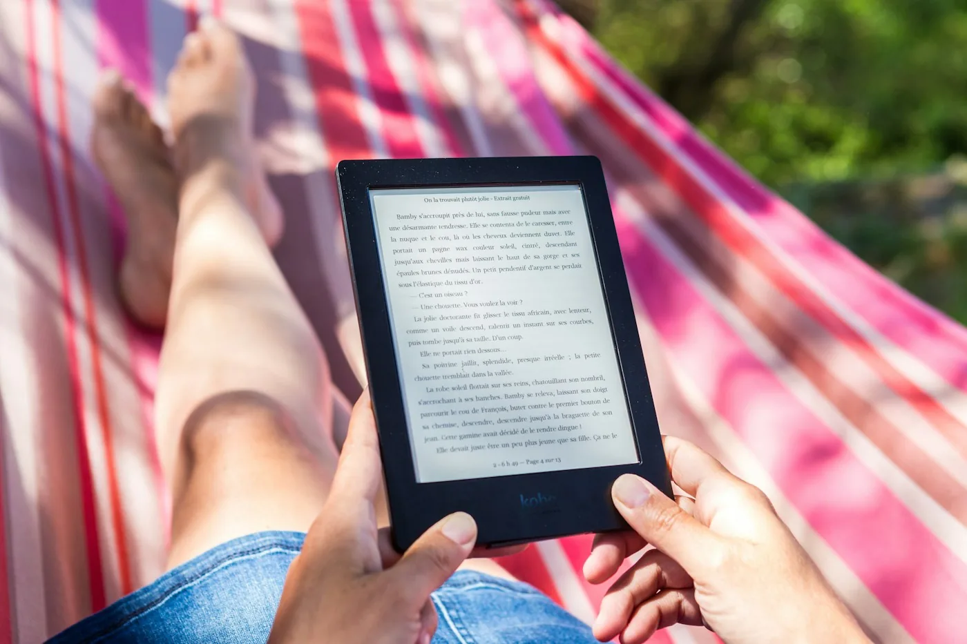 Manos sosteniendo un lector Kindle sobre una persona tumbada en una hamaca a rayas rojas y blancas en el exterior