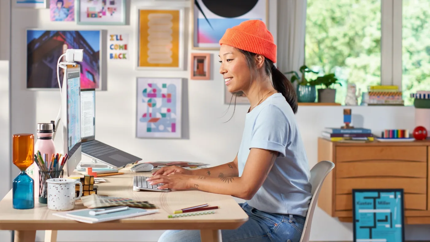 Woman in an orange beanie typing on a compact Logitech keyboard at a home desk with a laptop and artwork behind her