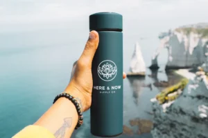 A hand holding a dark green bottle up against a calm sea with white chalk cliffs