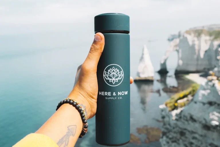 A hand holding a dark green bottle up against a calm sea with white chalk cliffs