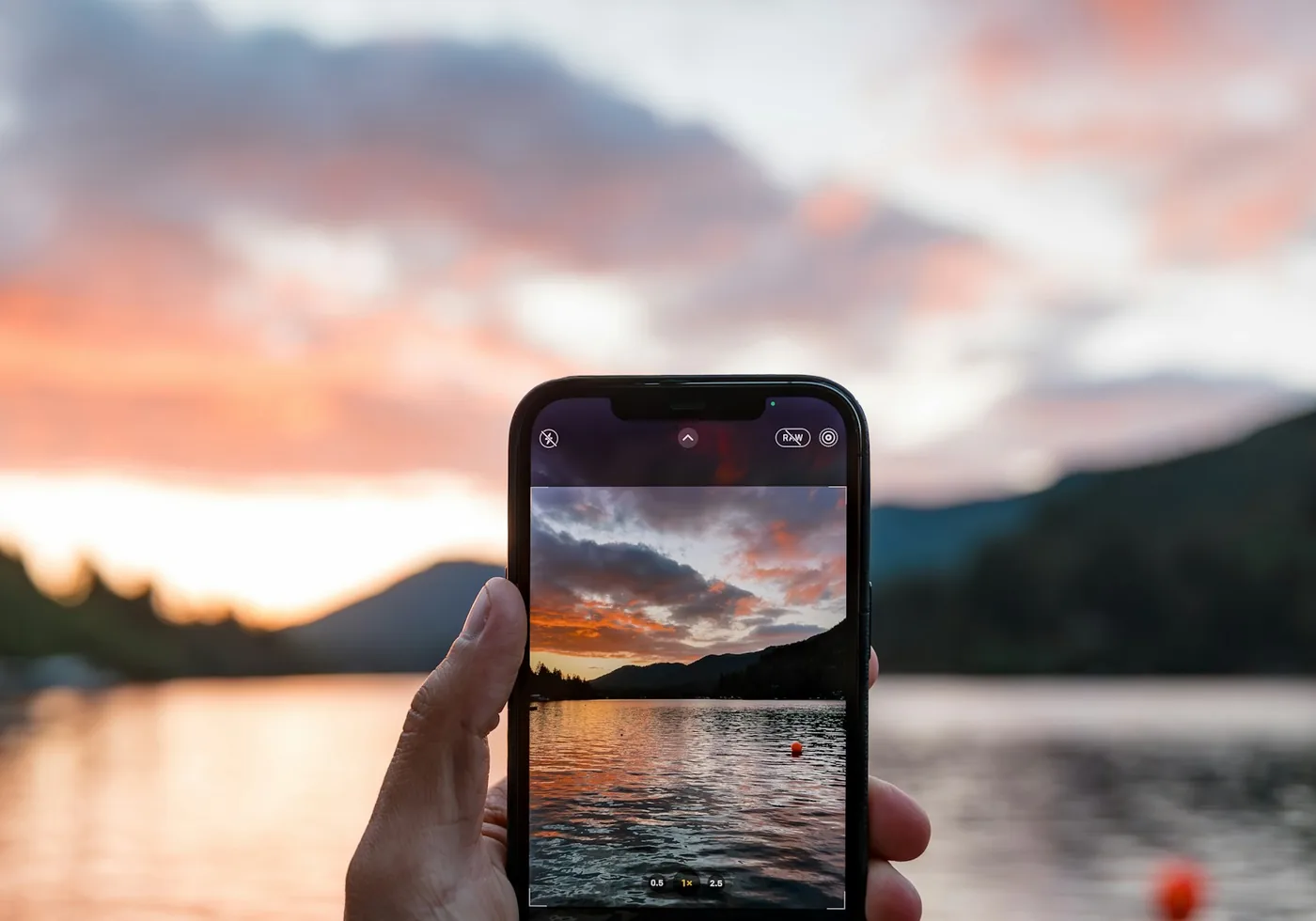 Hand hält ein Smartphone, das einen Sonnenuntergang über einem See und Bergen zeigt