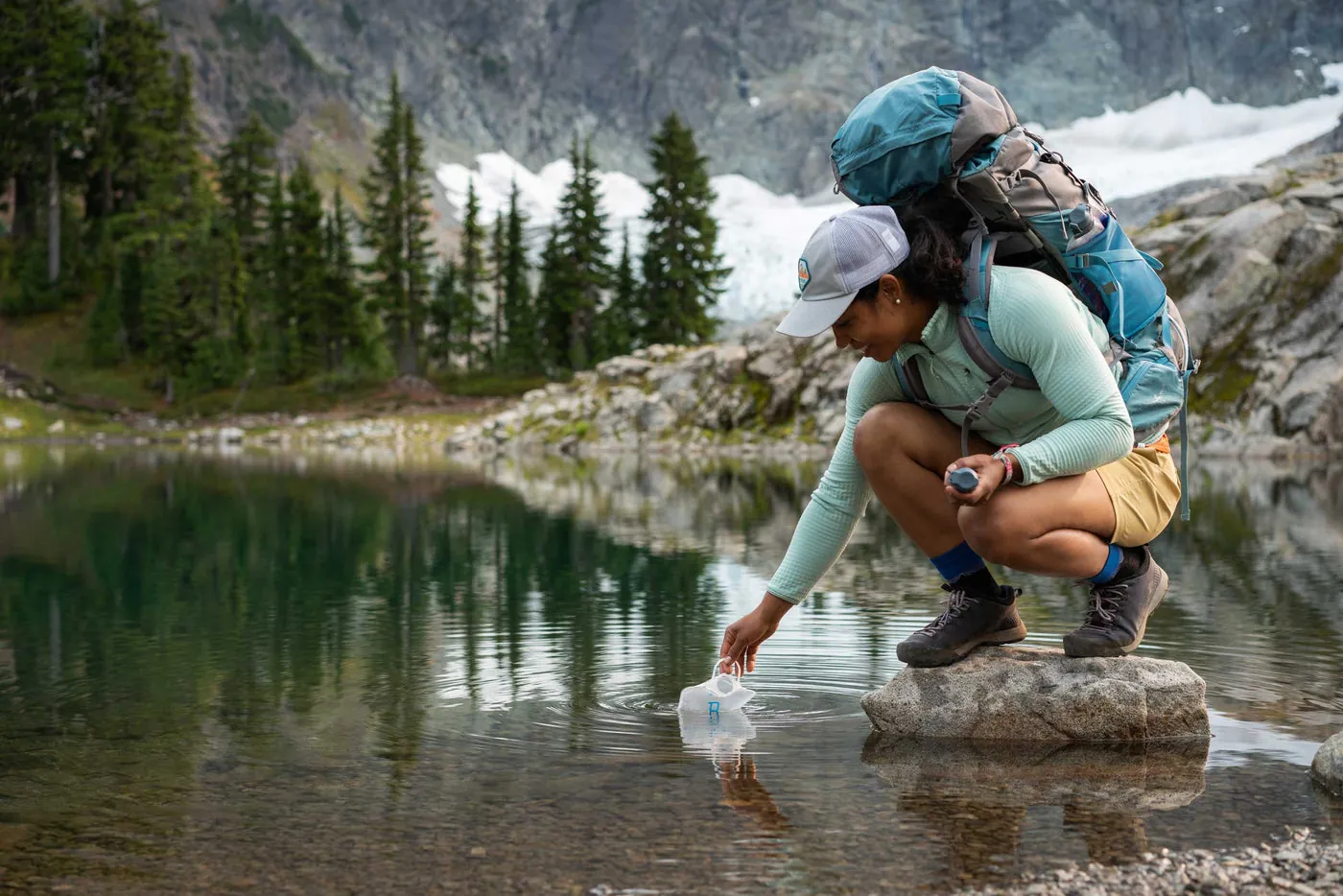 Wanderer mit türkisfarbenem Rucksack hockt auf einem Felsen und füllt ein Platypus-Reservoir aus einem Bergsee