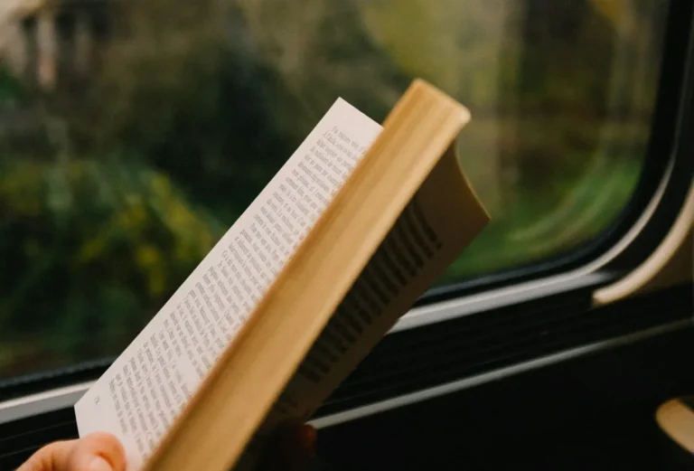 Hand holding an open paperback book by a train window with trees outside