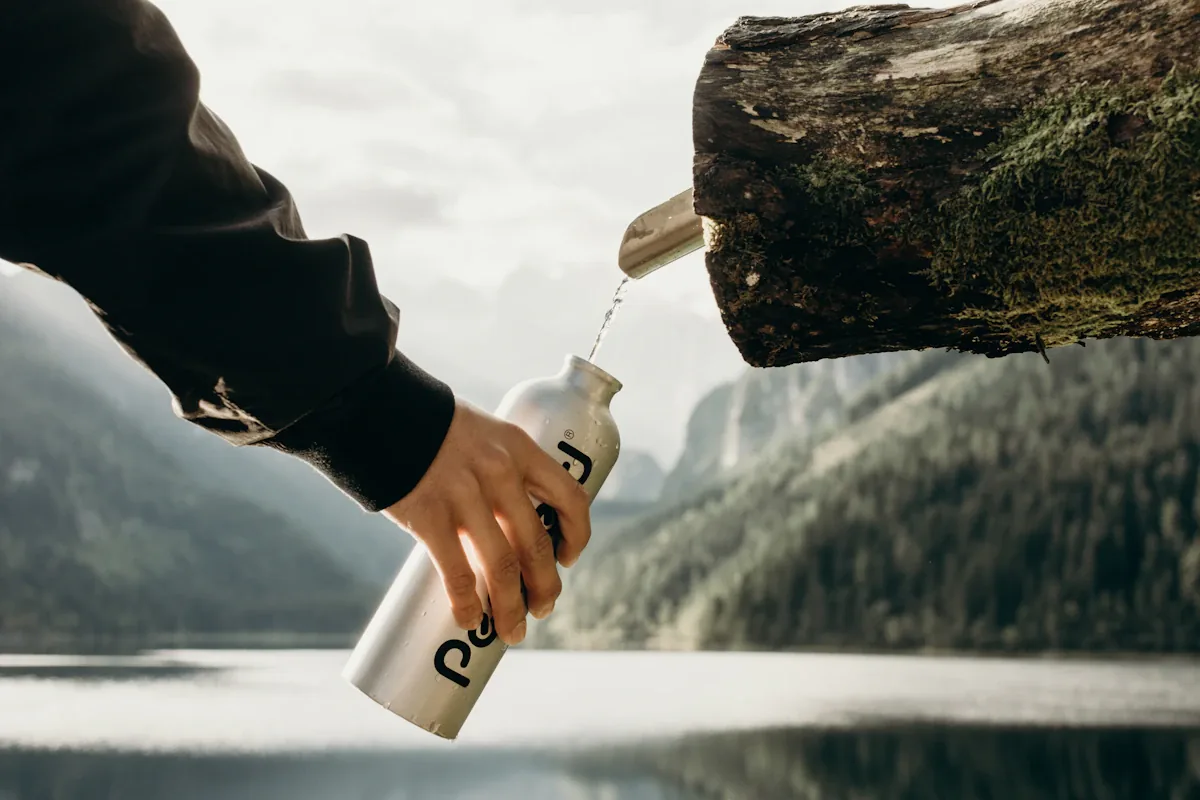 Een hand die een stalen fles bijvult uit een tuit op een boomstam boven een bergmeer