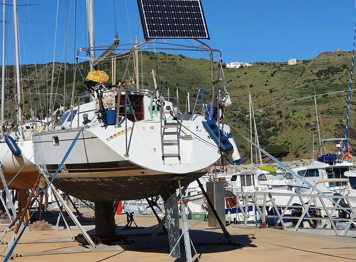 Segelboot im Hafen mit einem Solarpanel auf einem Ständer am Heck und einem grünen Hügel im Hintergrund