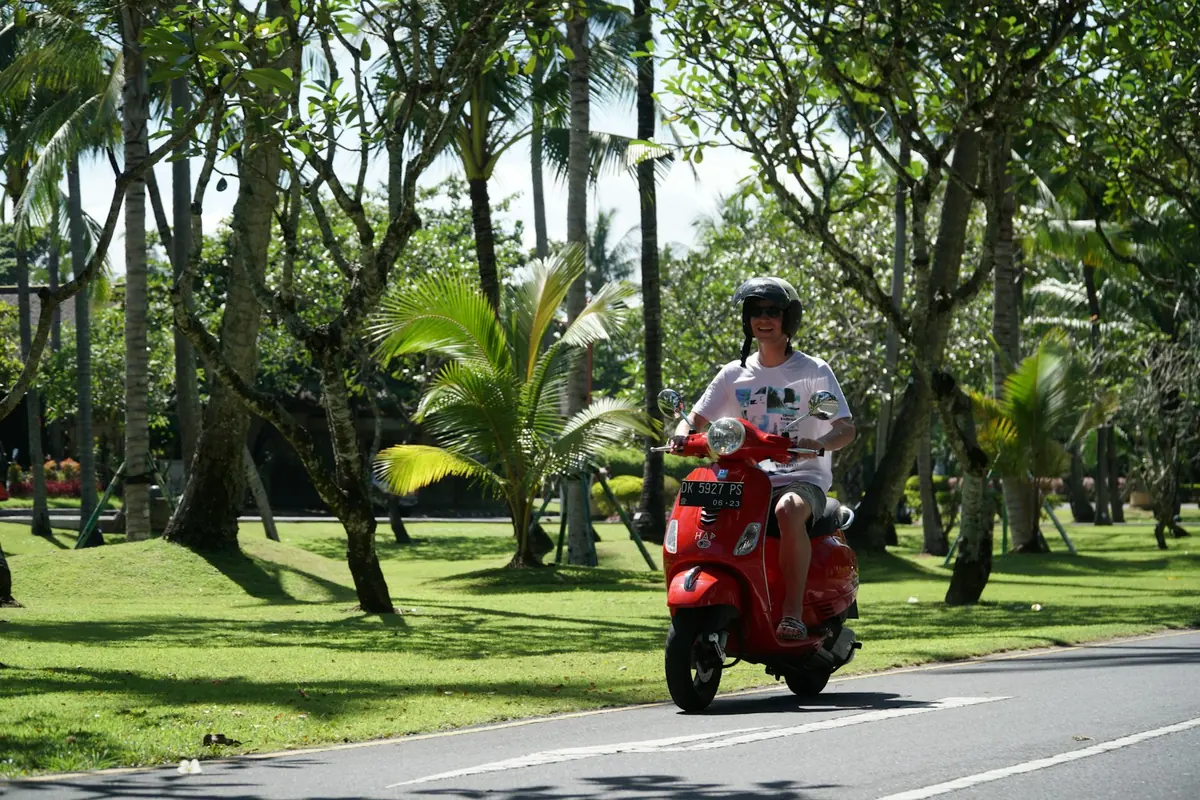 Rider with a white helmet on a red scooter on a palm-lined road, scooter rental checklist in Bali