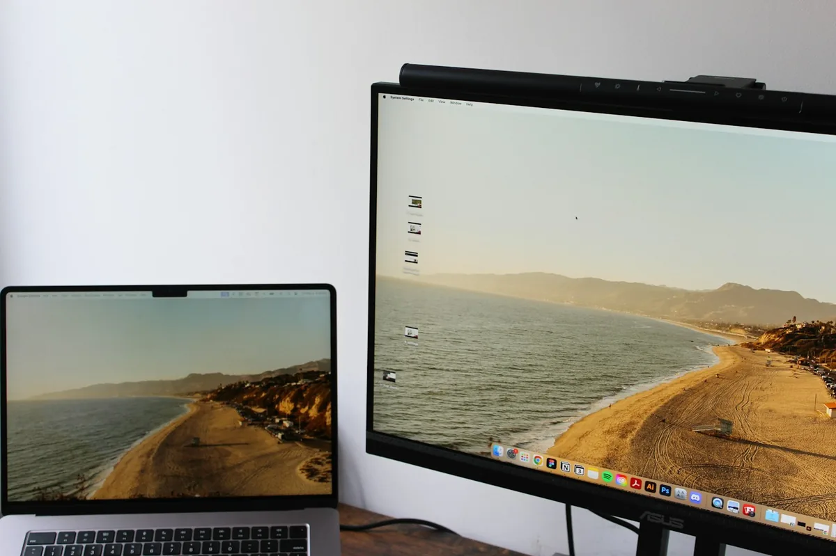 Laptop on a desk beside a larger monitor with the same beach wallpaper, close-up of the screen edge