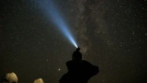 Silhouette of a man sitting on a rock at night with a headlamp beam pointing up into the starry sky