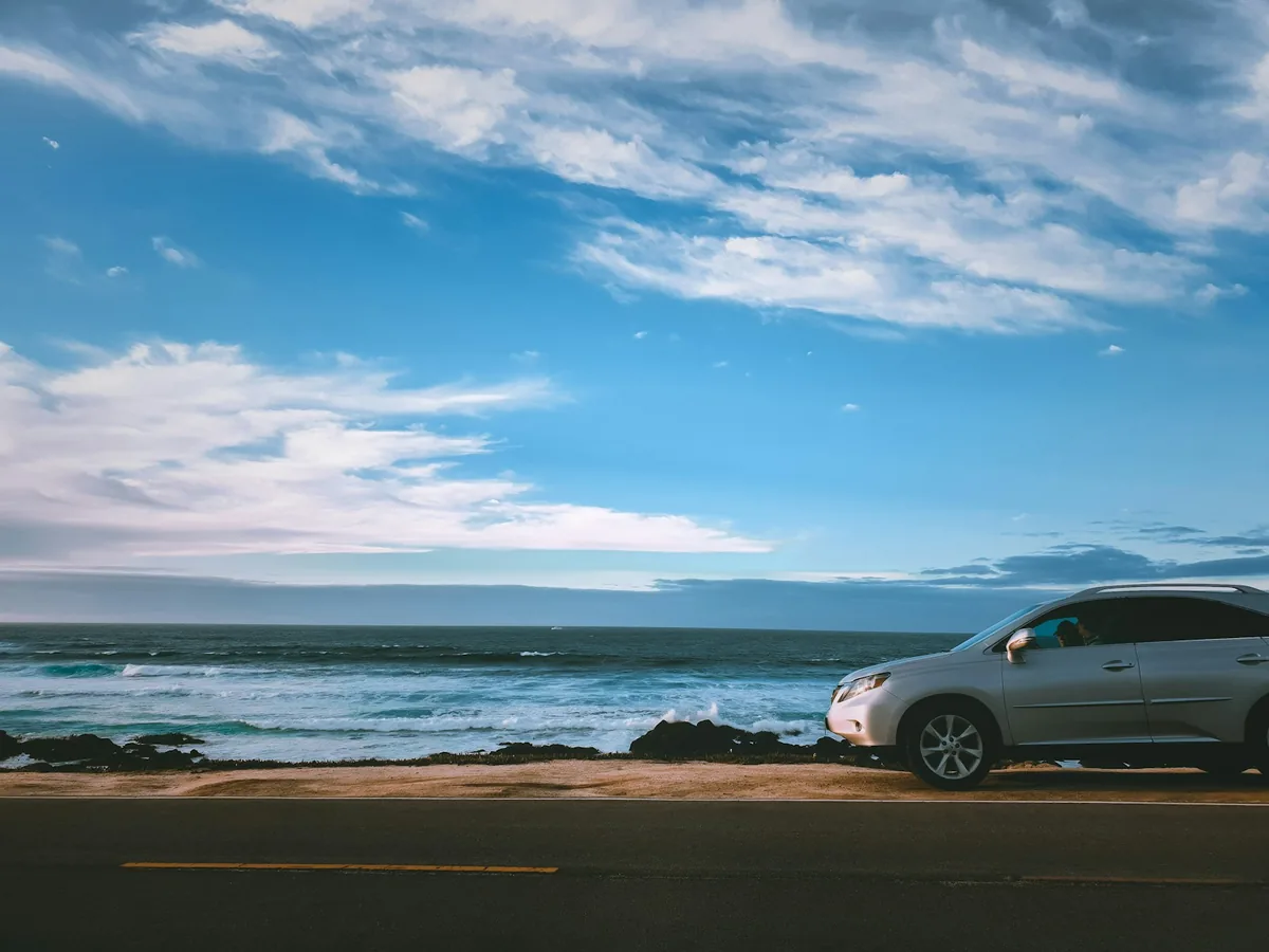 Silver car parked beside the sea under a blue sky with scattered clouds