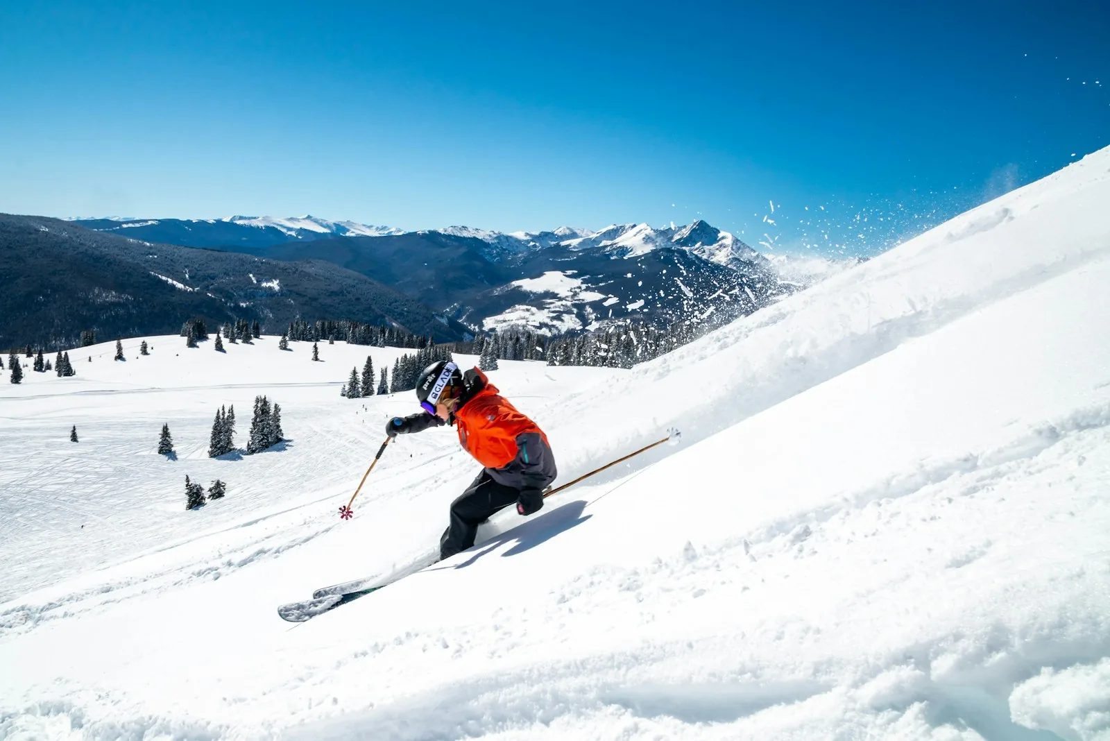 A skier in an orange jacket carving down a sunny slope with snow spray
