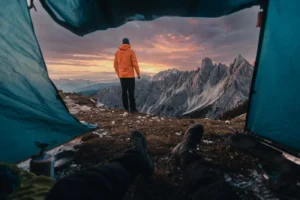 Hiker in an orange jacket seen from inside a tent, looking at a jagged mountain range at sunset