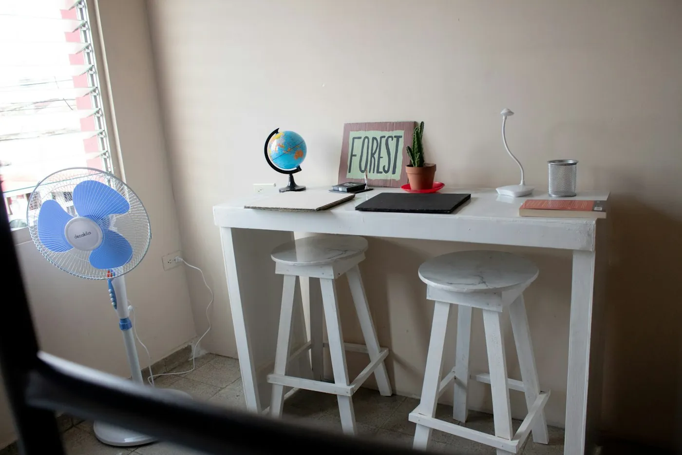 Small white desk with a globe, a plant, a notebook and two white stools beside a fan in a bright room
