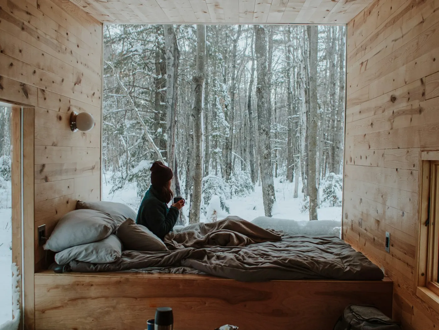 Person sitting on a bed in a small wooden cabin looking out of a large window at a snowy forest