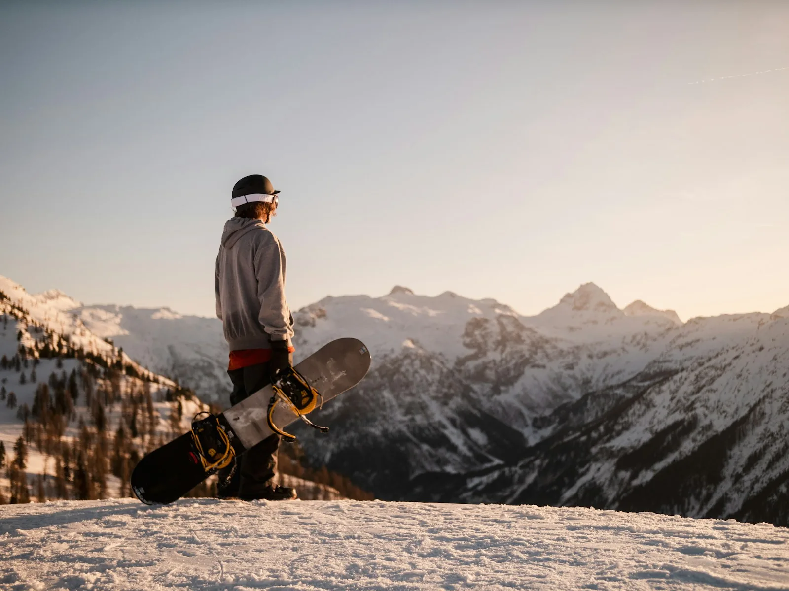 A snowboarder standing on a ridge at sunset with a board under his arm and mountains behind him