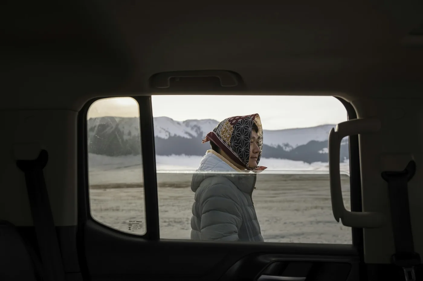 Woman in a white puffer jacket and a headscarf seen through a vehicle window with snowy mountains behind her