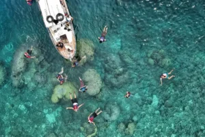 Swimmers snorkelling around a boat in clear turquoise water, seen from above