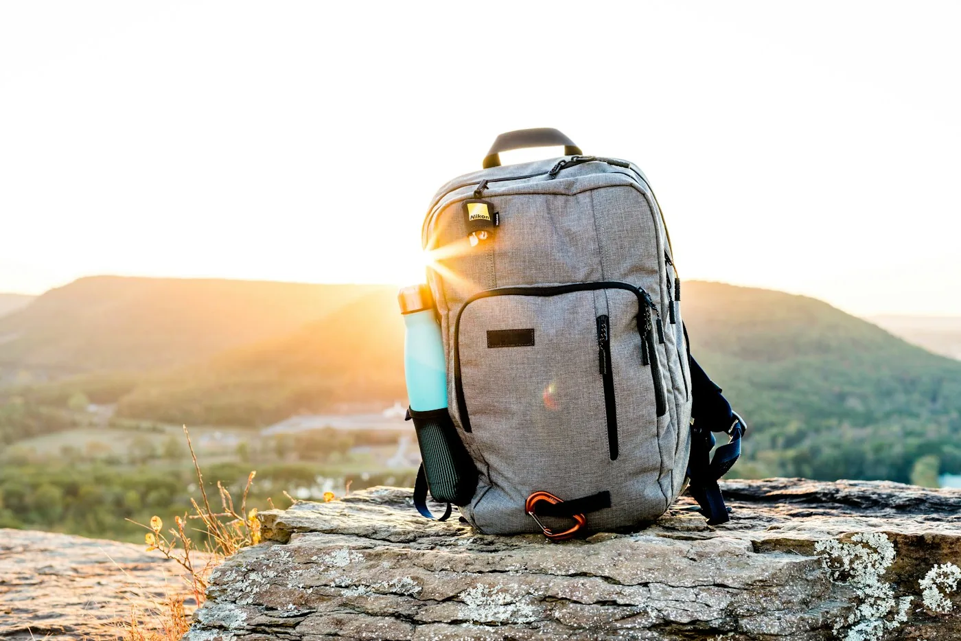 Grey and black backpack with a cyan tumbler on a rock at sunset with hills behind