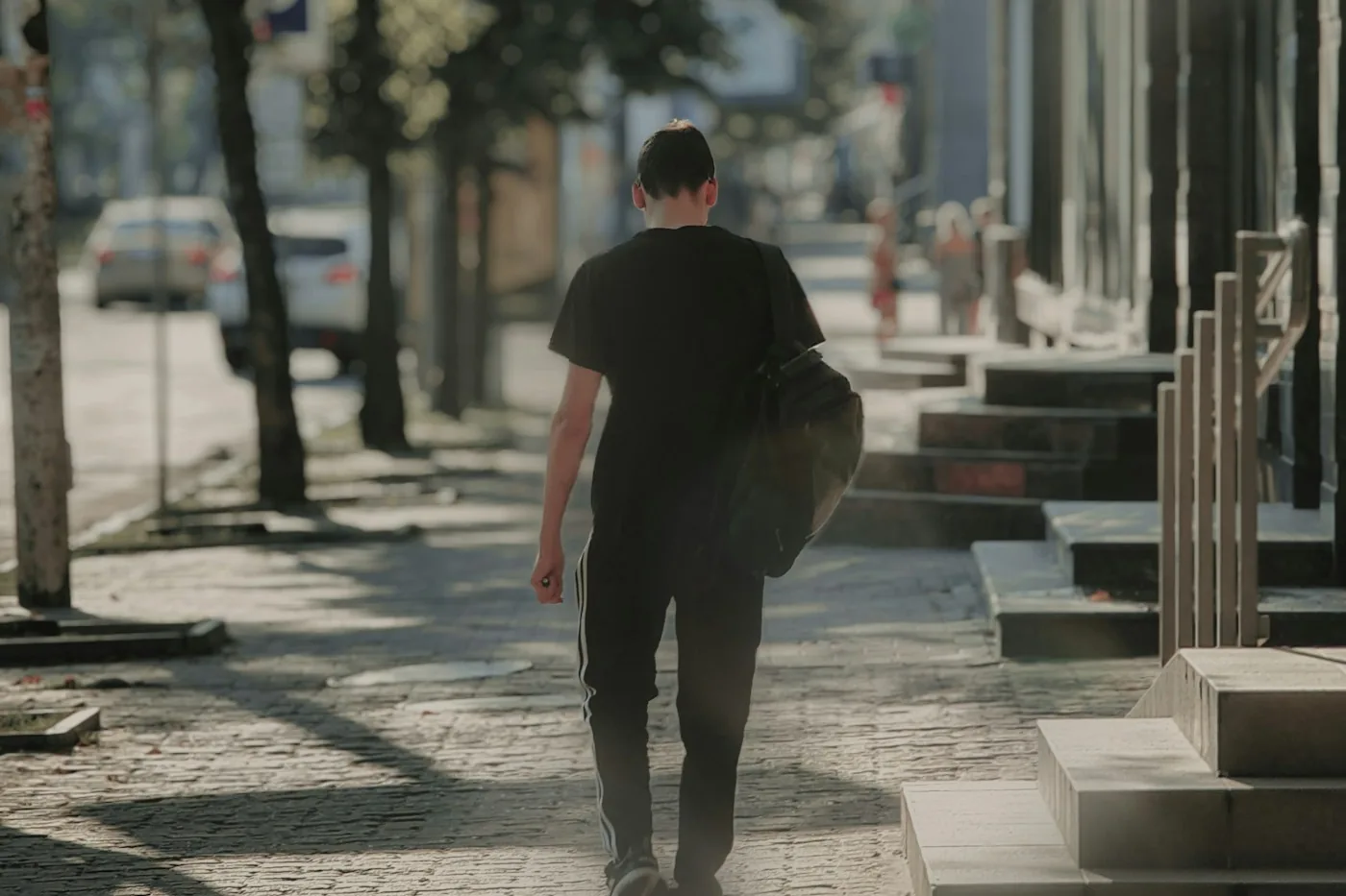 Man in a black hoodie with a backpack walking away down a sunlit city street