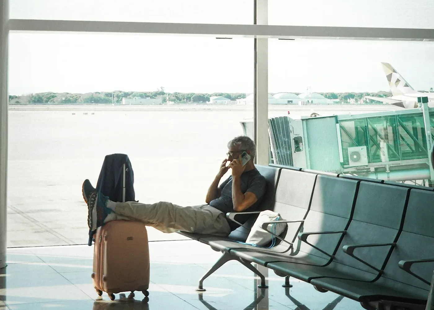 Man on a phone call in an airport seat with his feet up on the next seat and a suitcase beside him