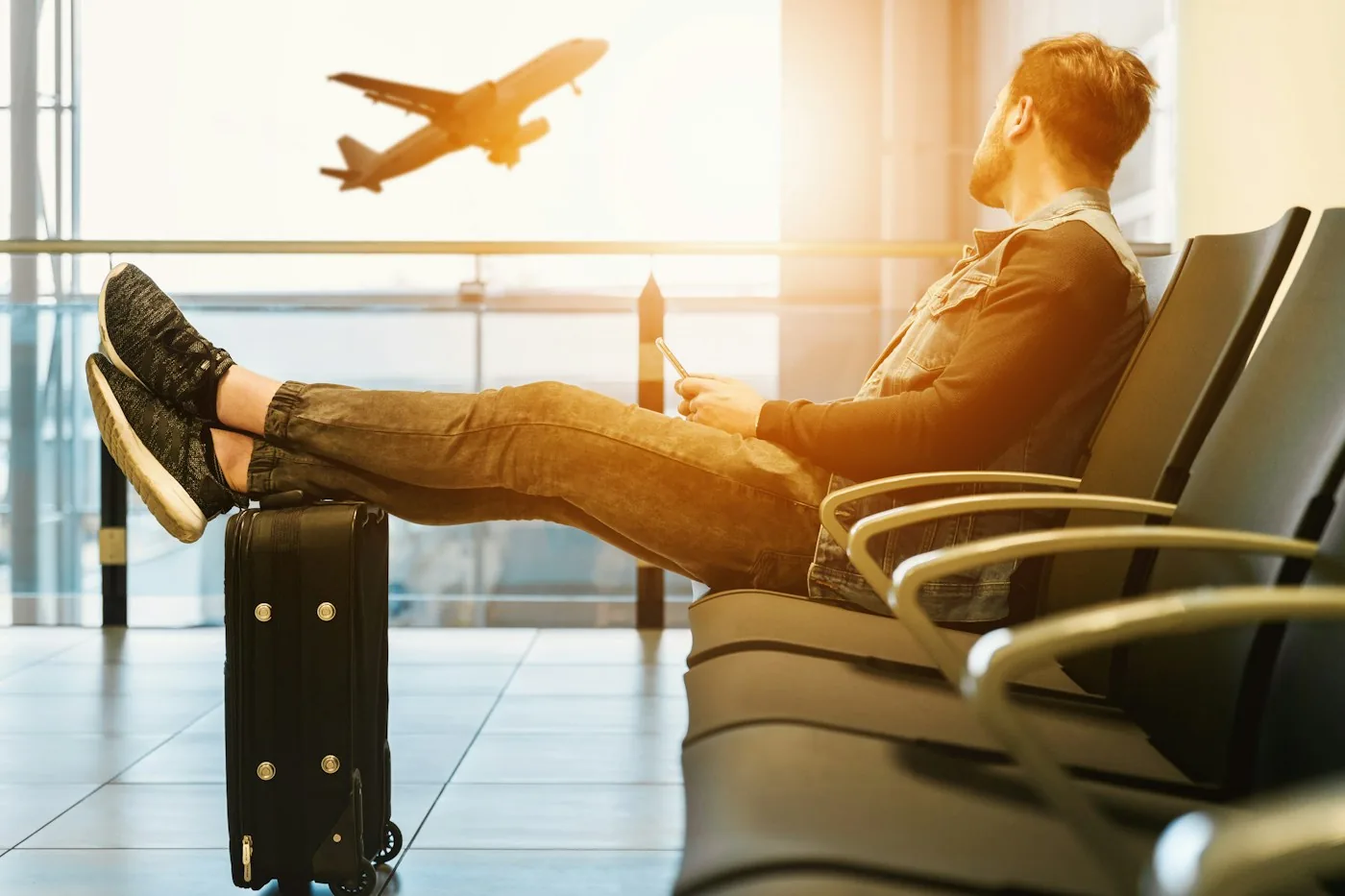 Traveler sitting in an airport lounge with feet on a suitcase, watching a plane take off