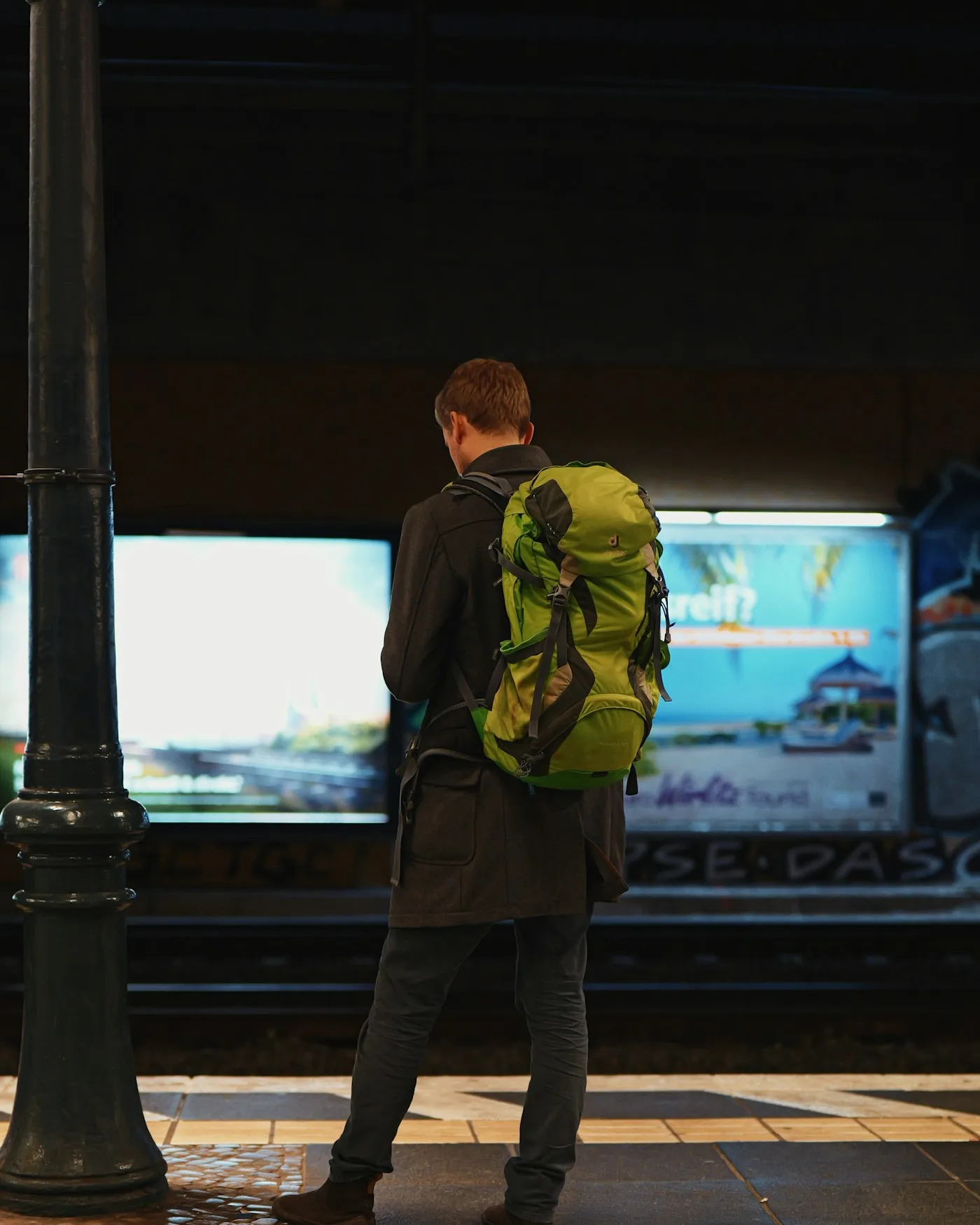 Viajero con mochila verde visto de espaldas, esperando en el andén de una estación