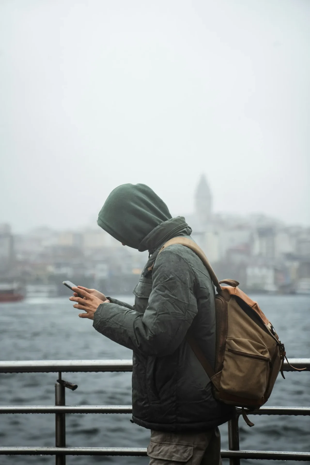 Ein Reisender mit Rucksack und Kapuze schaut auf sein Handy am Wasser