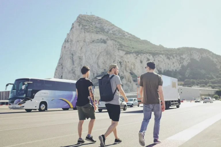 Three travelers with backpacks walking along a road near a coach, with a large rock behind them