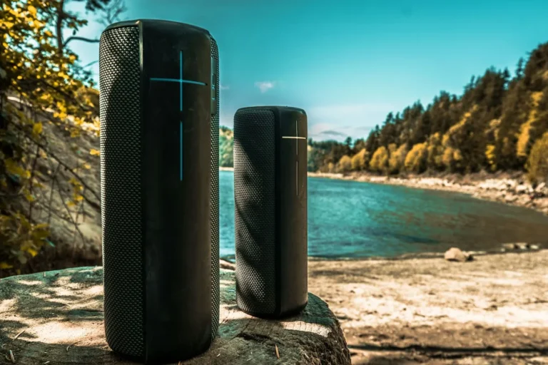 Two black waterproof Bluetooth speakers standing on a rock above a turquoise lake shore