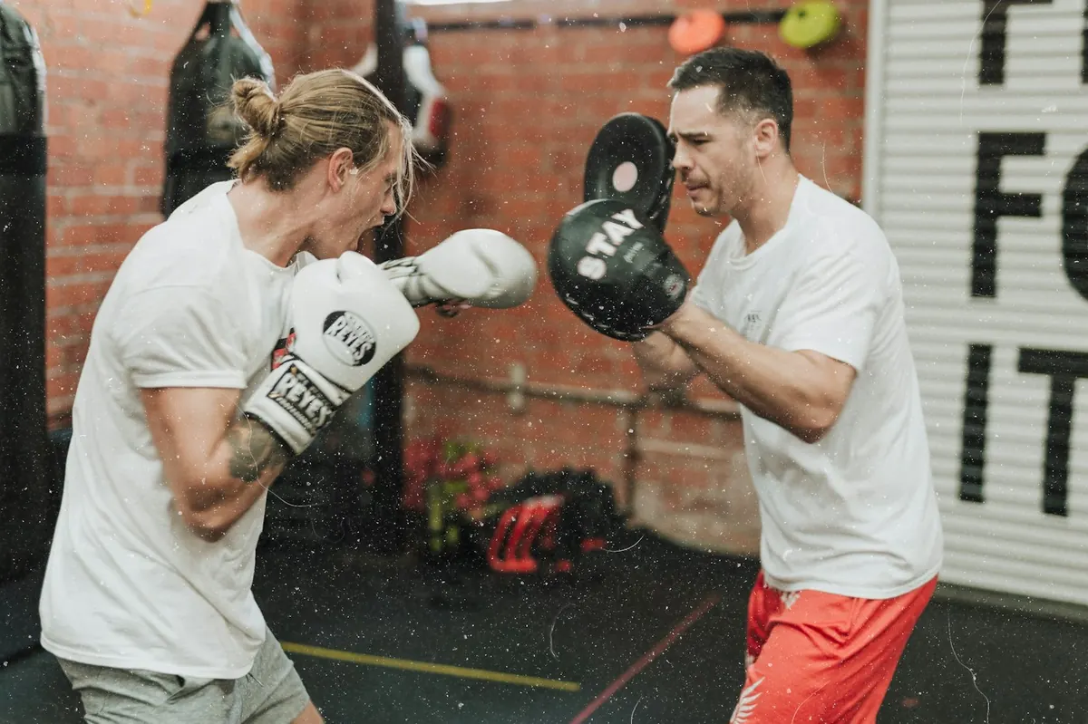 Boxer in a white t-shirt throwing a punch at focus pads held by a coach in a brick gym