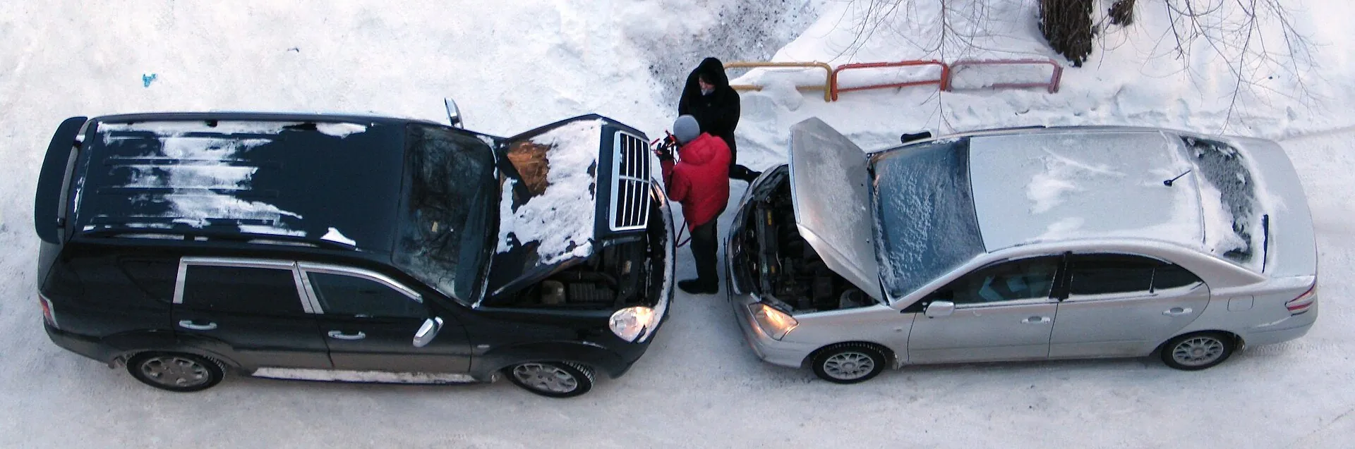 Two cars nose to nose with open hoods in the snow, seen from above, jump-starting with cables