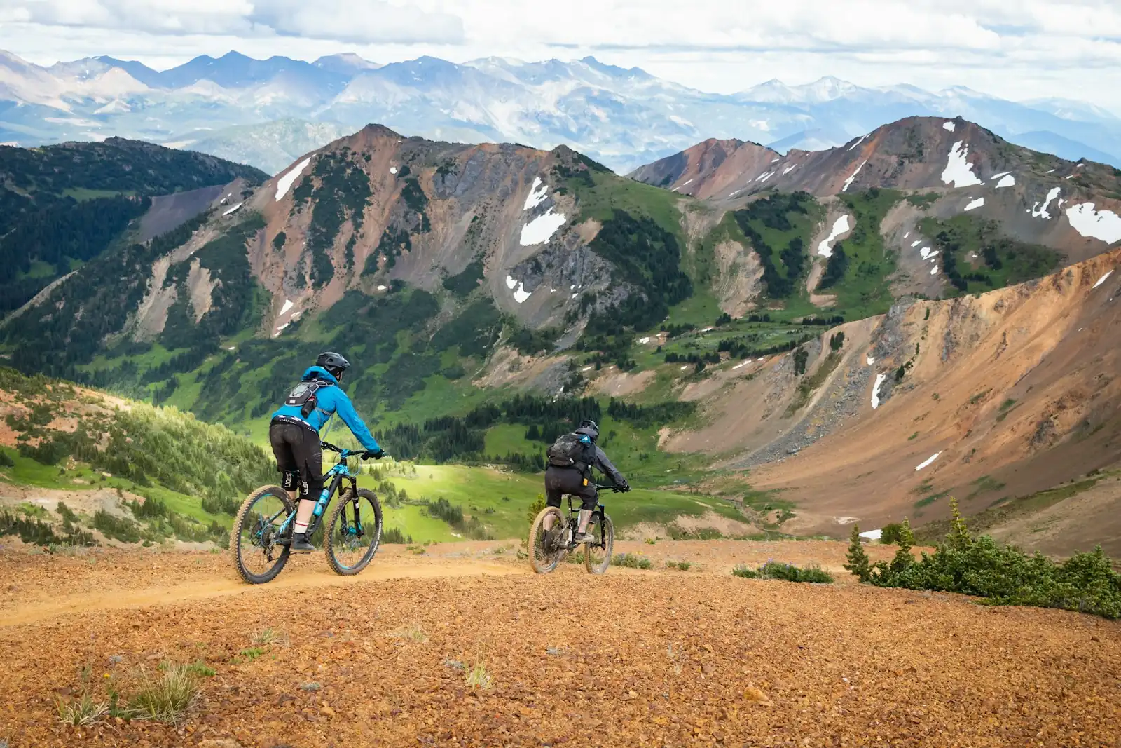 Dos ciclistas de montaña en una cresta de grava naranja sobre un valle verde y picos rocosos