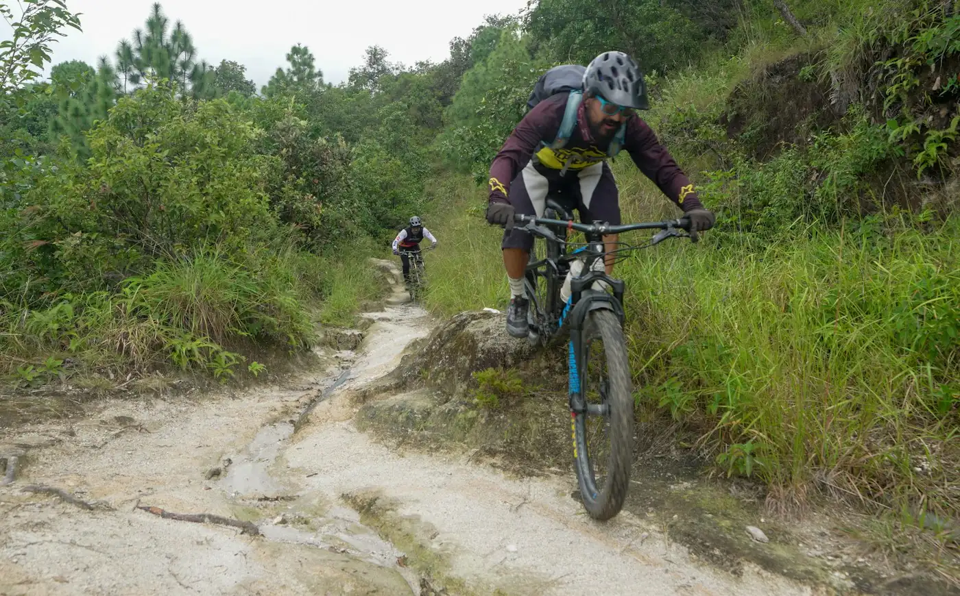 Dos ciclistas de montaña recorriendo un sendero rocoso entre matorrales de una colina verde