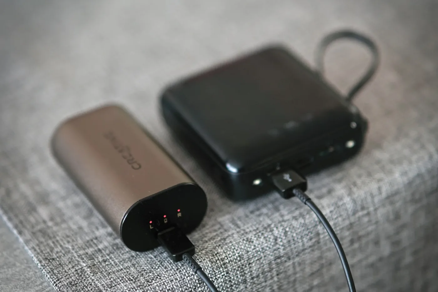 Zwei Powerbanks, eine bronzefarben und eine schwarz, laden auf einer grauen Stoffoberfläche
